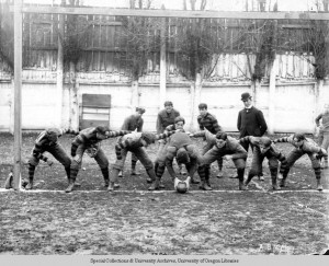 Black and white photo of the 1899 University of Oregon football team in a posed picture taken at Multnomah Field in Portland, showing the offense ready to snap the ball. 